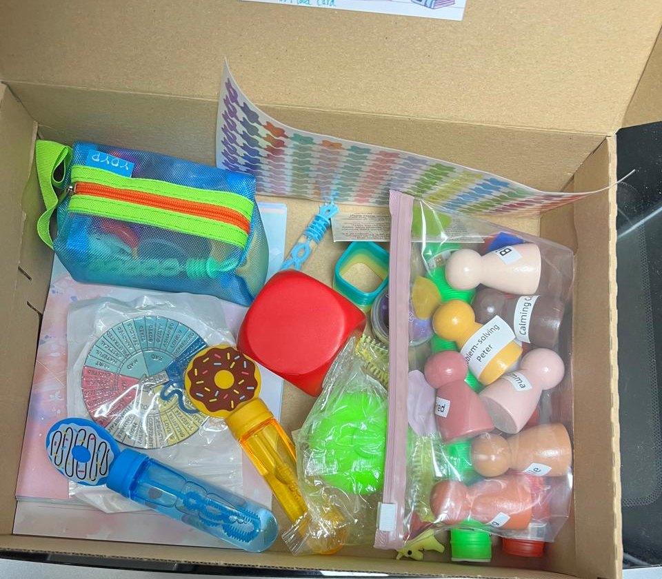 An open coaching toolkit box containing a feelings wheel, wooden figures, sensory items and bubbles.